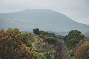 Obraz premium The historic Settle to Carlisle railway tracks pass under the stone Force Gill Aqueduct, with the distinct flat-topped plateau peak of Ingleborough rising into a misty, overcast Yorkshire sky.