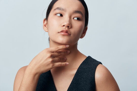 Young woman with long black hair wearing dark sleeveless top posing and touching her neck with thoughtful expression on light gray background in studio portrait.