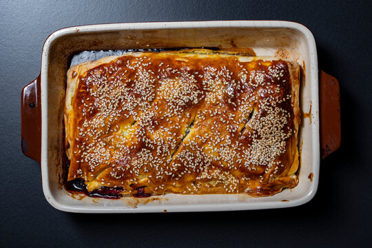 Freshly baked sweet berry pie with glossy egg wash and sesame seeds in ceramic baking pan, closeup. Crispy golden crust homemade pastry dessert on dark background, flat lay top view