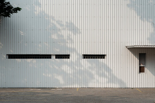 White corrugated metal factory facade with narrow windows, door awning, and tree leaf shadows.