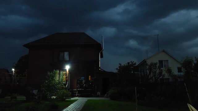 Exterior of residential house at night with bright illumination. Dusk view of cottage with glowing porch light under cloudy sky. Garden path in dark evening.