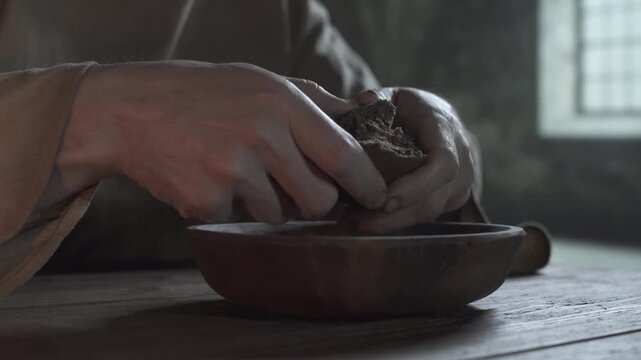 Hands of a medieval prisoner tearing a rye bread chunk over a wooden bowl inside a gloomy jail cell