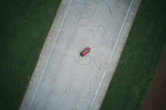 Aerial top-down view of a red car drifting and performing donuts on a concrete runway, leaving dramatic swirling tire marks