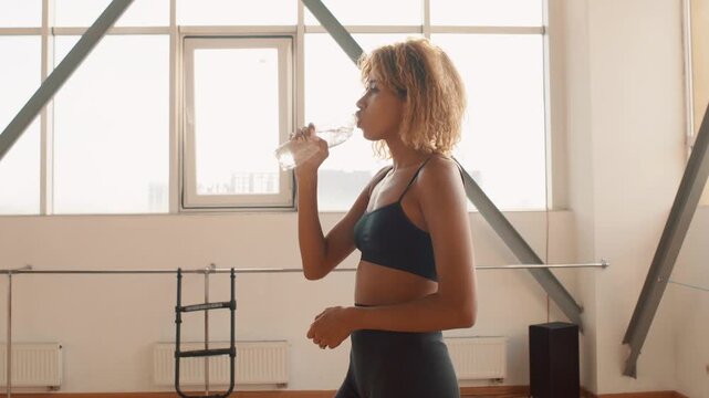 Fit Afro young woman drinks water after an indoor workout in bright sports gym, coolin down and rehydrating. Casual athletic wear, natural light and urban window backdrop emphasize health and recovery