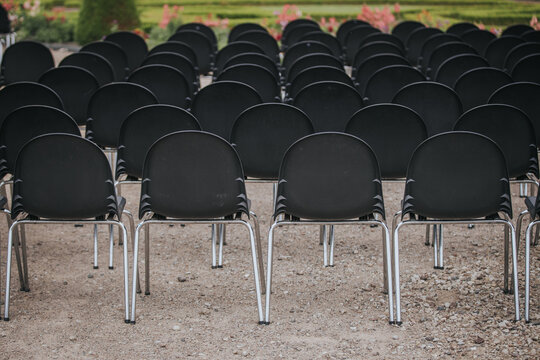 Rows of black plastic chairs with chrome legs arranged neatly on gravel ground in an outdoor setting, surrounded by green grass and flower beds
