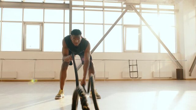 Young African American athlete intensely performs battle rope exercises in a spacious, modern gym bathed in sunlight. This dynamic workout emphasizes focus, power, and dedication Front View 4K UHD