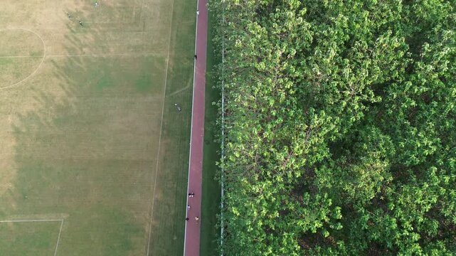 Top-down aerial drone view of a football field beside a running track and lush green trees. People are visible exercising and playing sports in an outdoor recreational area, creating a balanced compos
