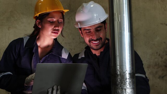 Two industrial engineer workers in safety suit and helmet operating machinery together at manufacturing metal factory during night shift. Woman staff uses laptop, discuses with foreman while working.