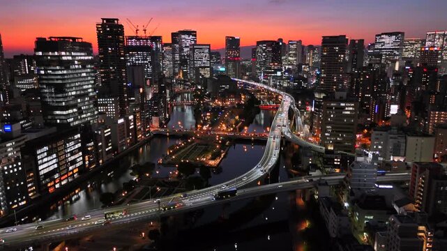 Beautiful static aerial drone view of modern Osaka city skyline at colorful sunset. Illuminated downtown skyscrapers, busy highway traffic and river bridges during twilight in Kansai region Japan