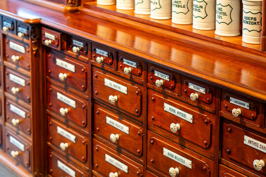 Close-up of an antique wooden pharmacy cabinet with numerous drawers featuring white ceramic knobs and labels with herb names, and a row of white porcelain jars placed on the countertop.