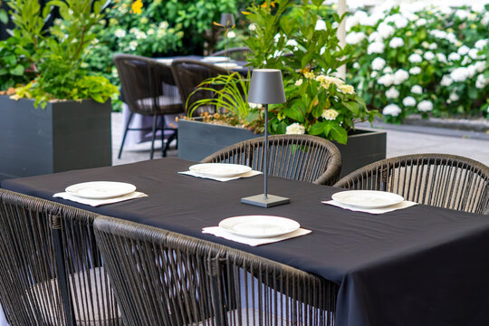 Close-up of an elegant caf&eacute; table covered with a black tablecloth, featuring white plates and a minimalist table lamp, surrounded by dark rattan chairs on a terrace filled with greenery and blooming w