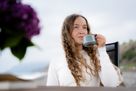 portrait of curly-haired woman enjoying a warm drink by the seaside and peacefully looking into the distance. Cozy coastal lifestyle, slow living and mindful relaxation concept.