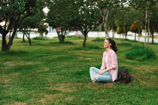 A smiling woman sits on green grass in a sunlit park, enjoying a peaceful moment. A lifestyle image highlighting outdoor serenity and everyday calm.