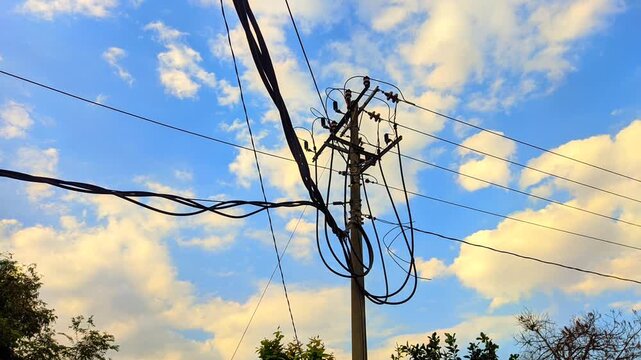 Time lapse of concrete utility pole with messy electric power lines and cables against a moving cloudy blue sky during golden hour evening.