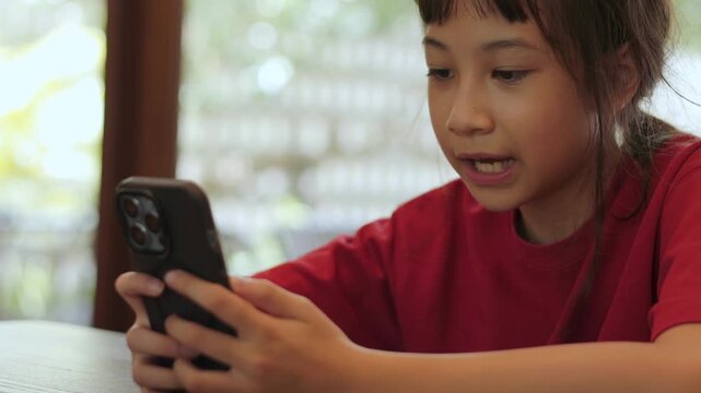 Asian kid using a smartphone at restaurant. Cute Thai girl excitedly watching video or playing games. Shot with selective focus and beautiful copy space for text. Childhood digital lifestyle concept.