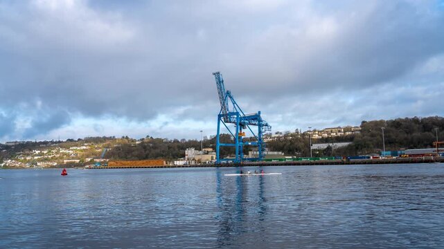 A large blue gantry crane stands over a port, with a pile of logs and shipping containers visible on the dock. 