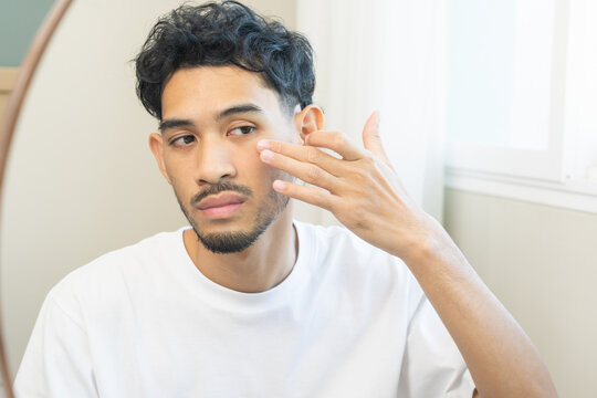 Guy skin care beauty concept, worry asian young man touching his skin while looking in the mirror, stressed male with problem presenting on face. Health care from acne problem, dry skin, treatment.