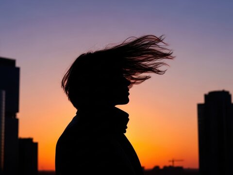 Silhouette of woman with windblown hair against colorful sunset sky and city skyline