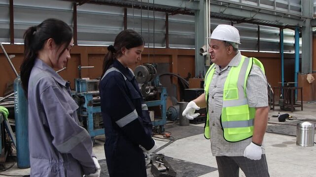 Grumpy elderly engineer supervisor with safety vest and white helmet scolding two Asian female workers. Hot-tempered senior foreman shouting at industrial staff in anger at manufacturing metal factory