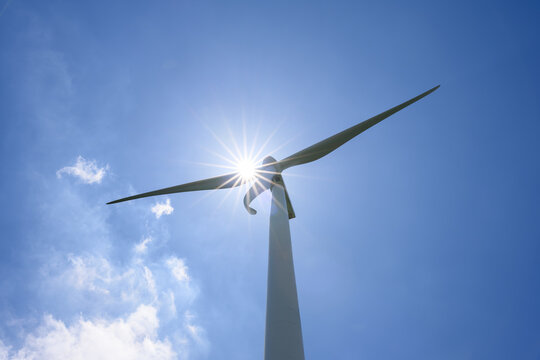 Wind Turbine Reaching Into Bright Blue Sky, Sun Flare Bursting Between Blades, Symbol of Clean Renewable Energy