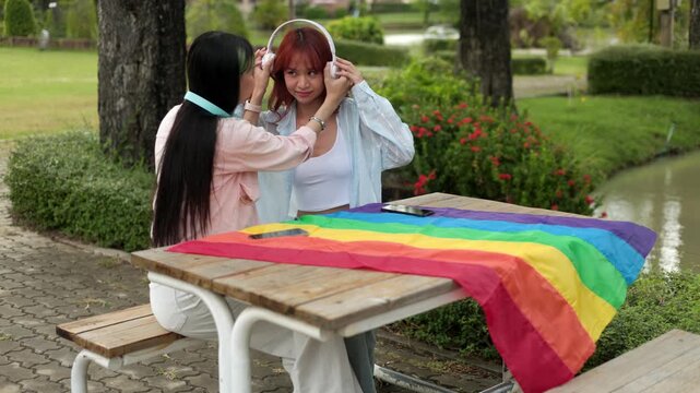 Two beautiful Asian women sitting together at a wooden table in the park, adjusting headphones to listen to music, sharing a happy romantic moment during a pride month celebration.