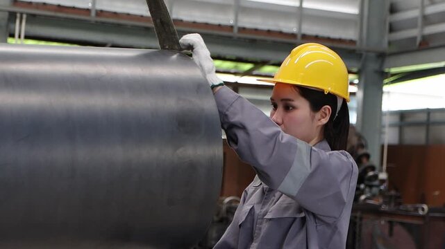 Industrial Asian female worker in safety suit and helmet measuring a large metal pipe being lifted in the air by machine at manufacturing factory. Staff checks quality of products on production line.
