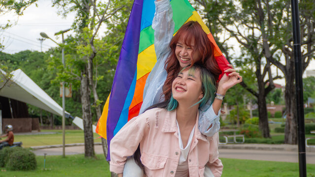 Happy Asian young LGBTQ lesbian couple piggyback with rainbow pride flag at park. Gen Z female lovers smiling, enjoying romantic relationships, freedom, gender diversity, equality.