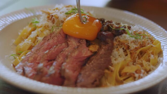 Close up of piercing a runny egg yolk over a delicious pasta dish with sliced medium-rare beef steak and grated cheese. Appetizing gourmet food and mouth-watering culinary dining concept.