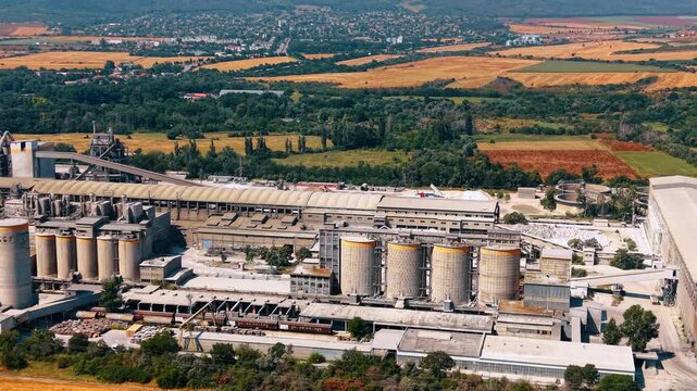 Cement factory silos and processing buildings from above. Oblique aerial view of cylindrical storage tanks, rail wagons, conveyors and patchwork farmland in daylight.