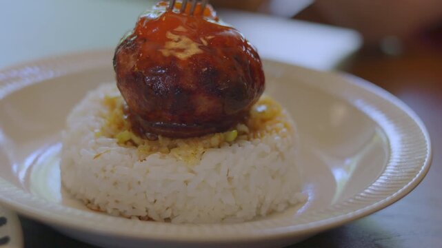 Close up of fork pressing a juicy hamburg steak on top of white rice. Savory meat juices and fat oozing out. Mouth-watering gourmet meal shot with selective focus. Delicious culinary dining concept.