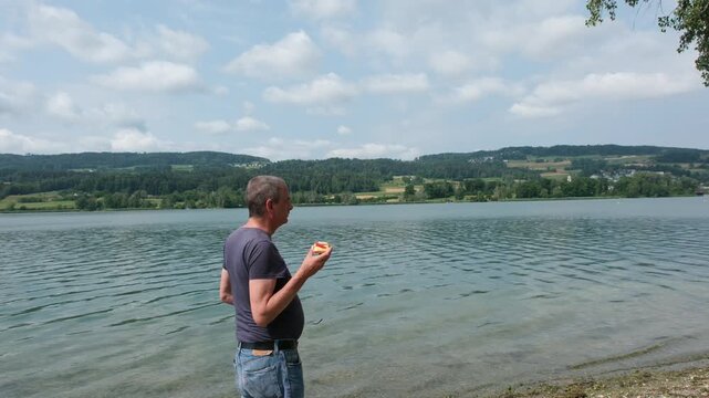 Hunger und Lust auf schnelles Essen im Sommer in der Natur, Mann isst in der Freizeit zwischendurch ein kalorienhaltiges Brot am See als willkommene Zwischenverpflegung