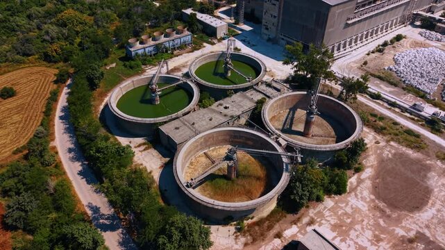 Circular settling basins at cement factory. High angle view of four concrete clarifiers with central rotating arms, green water and dry interiors beside trees.