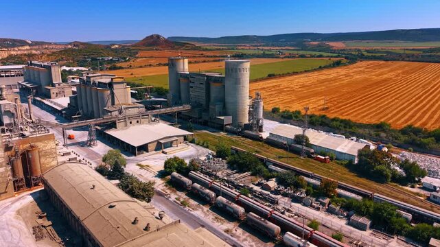 Cement silos beside railway and harvested fields. Aerial panorama of storage towers, factory buildings, freight wagons and orange crop rows across rolling farmland.