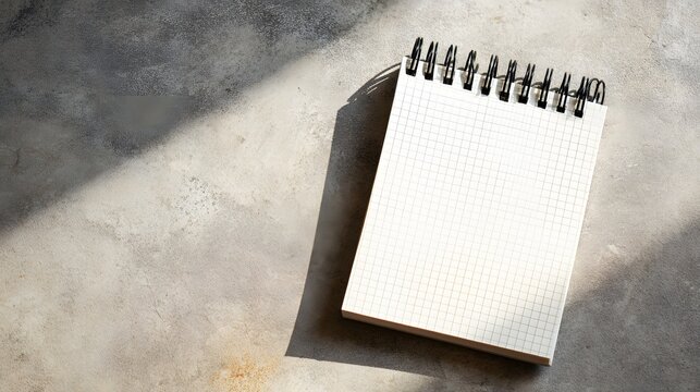 Top view of blank spiral grid notepad over textured stone surface with hard window shadow play backdrop.