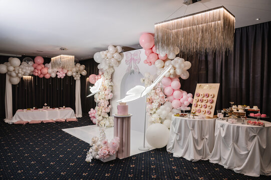 Elegant birthday party decoration in a restaurant for a festive reception, featuring a balloon arch, floral arrangements, glowing butterfly decor, candy bar, donut bar, and birthday cake.