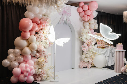 Elegant birthday party photo zone in a restaurant for a festive reception, featuring a white arch, pastel pink and white balloon garland, floral arrangements, glowing butterfly decor, a birthday cake.