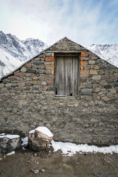Rustic stone building with weathered wooden window against snowy Mount Kazbek backdrop in Stepantsminda, Georgia.