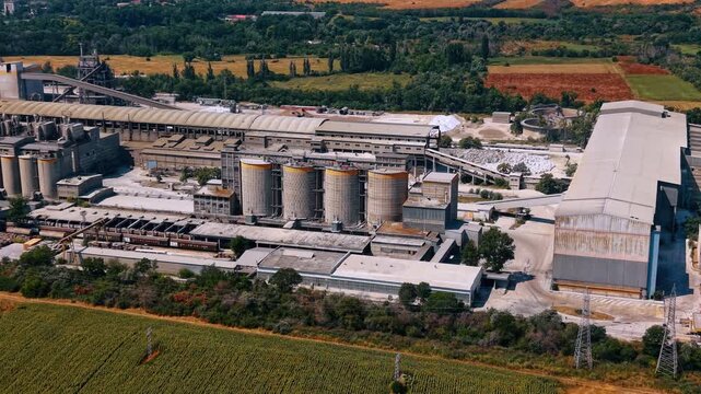 Cement silos and industrial halls beside crop field. Oblique aerial view of cylindrical storage tanks, rail wagons, conveyors, warehouses and sunflower rows.