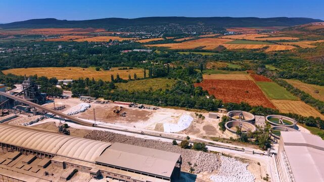 Cement plant material yard beside patchwork farmland. Aerial panorama of conveyors, stockpiles, circular basins, factory roofs, crop fields and a distant town.