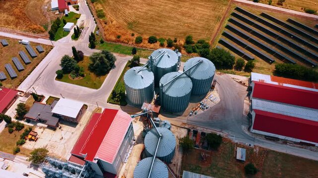Grain storage silos beside solar panel arrays. High angle aerial view of metal tanks with conical roofs, conveyors, red roof buildings, roads and photovoltaic rows.