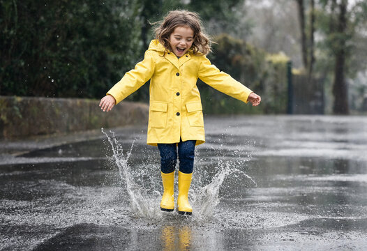 Happy young girl in a yellow raincoat jumping in a puddle on a rainy day outdoors in city