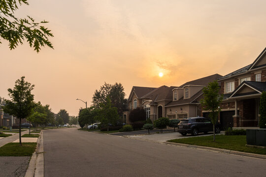 High-angle shot of a quiet suburban street with modern brick houses, set against an eerie orange and hazy sky caused by wildfire smoke obscuring the evening sun.