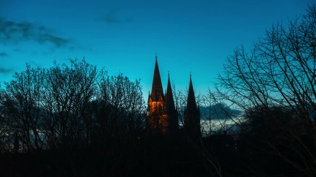 The illuminated spires of a church stand out against the twilight sky, framed by the bare branches of trees.