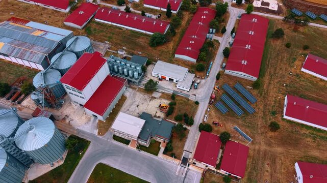 Farm processing hub with silos and solar panels. Oblique aerial view of red roof structures, storage tanks, machinery, roads and dry grass around the site.