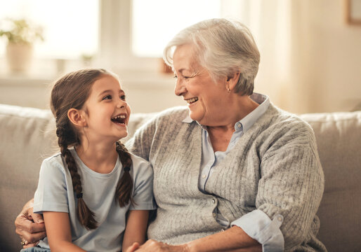 Cute school-age girl spending quality time with her elderly grandmother at home, multigenerational family sitting together on a cozy couch, laughing and enjoying a warm, trustworthy conversation.