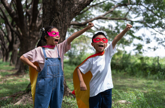 Two excited Asian children in superhero capes and masks shout loudly with fists raised in a nature park. The boy and girl show strong empowerment, brave teamwork, winning energy, and childhood fun.