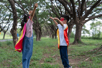 Obraz premium Two Asian children in homemade superhero capes and masks stretch their arms pretending to fly in a green park. The boy and girl play together, showing active imagination, victory, and fun.