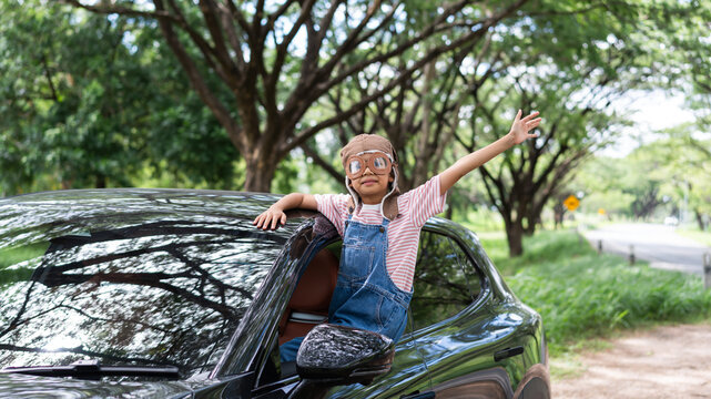 A happy Asian girl wearing an aviator hat and goggles leans out of a black car window with her arm raised. This road trip scene represents freedom, travel, and childhood imagination in nature.
