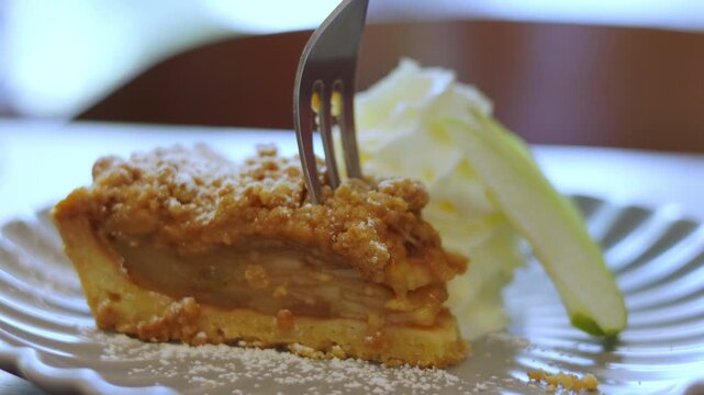 Close up of fork cutting into a delicious slice of apple crumble pie dusted with powdered sugar. Homemade sweet dessert with whipped cream. Shot with selective focus. Bakery pastry concept.