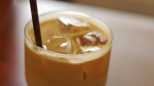Close up of a straw stirring refreshing iced coffee in a glass. Cold milk latte with ice cubes in natural light. Shot with selective focus. Morning cafe beverage and summer cooling drink concept.